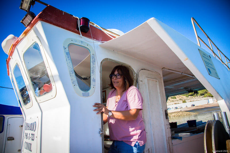 Mujer de pie en la cabina de un barco de pesca, saliendo por la puerta con una expresión sonriente. Lleva una camiseta rosa y gafas, y el barco está anclado en un puerto o cerca de la costa, con el cielo despejado de fondo.