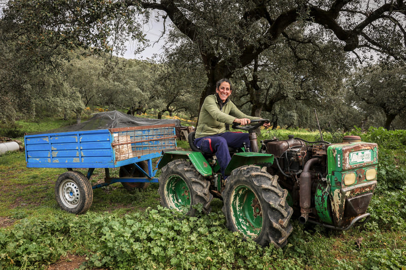 Mujer sonriente sentada al volante de un tractor verde con un remolque azul, en un entorno rural rodeado de árboles y vegetación.
